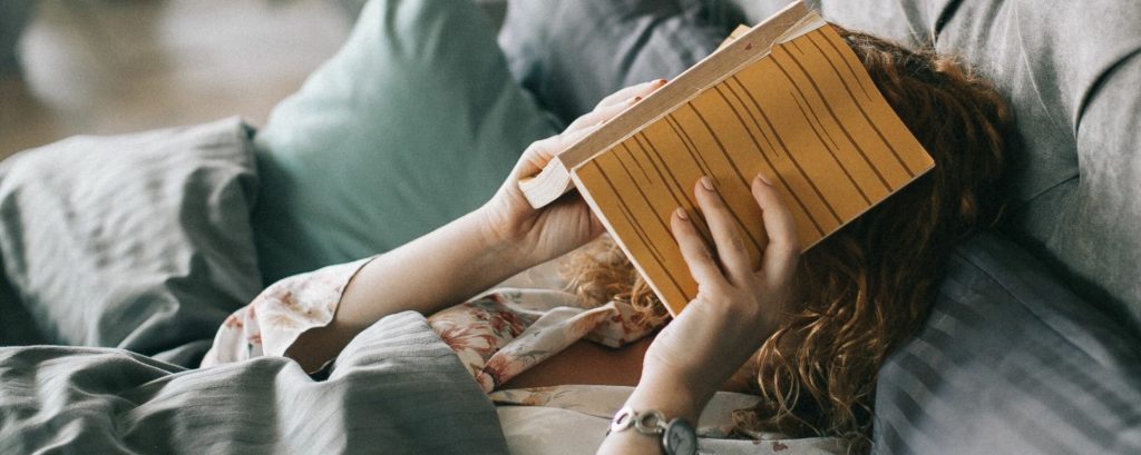 woman lying in bed hiding her face behind a book