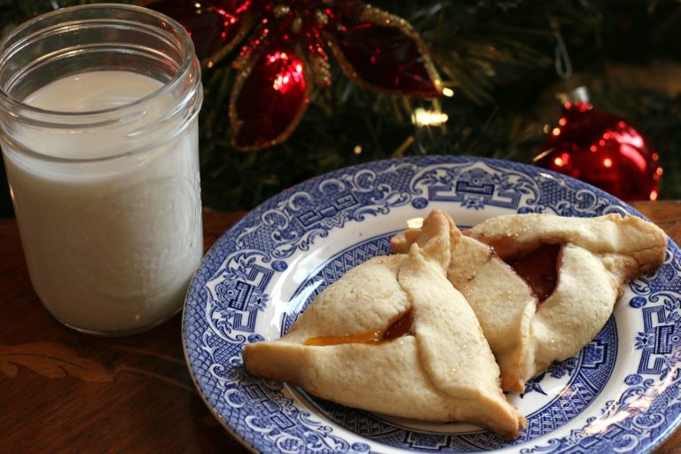 Two Hamantaschen cookies on a blue and white plate with a glass of milk
