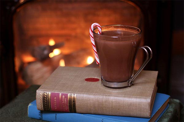 cup of cocoa with candy cane garnish on top of a pile of books in front of a lit fireplace