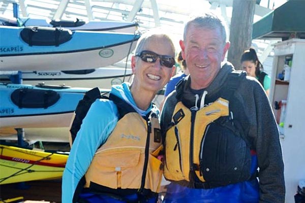 Smiling couple wearing life jackets in front of a rack of kayaks