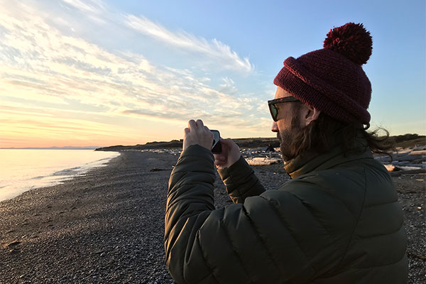 Man wearing a winter coat and hat on a rocky beach taking a picture with a cellphone