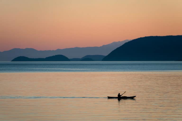 Profile of kayaker moving across still water at sunset