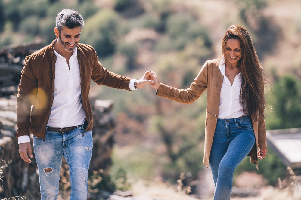smiling couple wearing white button down shirts, jeans, and tan jackets, holding hands